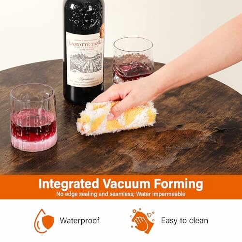 Hand cleaning a wooden table with spilled red wine and a bottle in view.