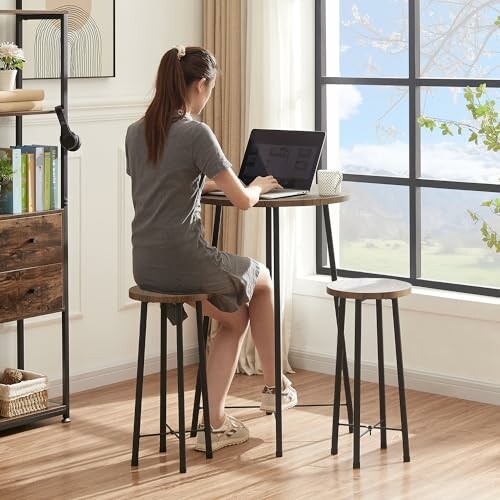 Woman sitting at a small table using a laptop in a bright room with large windows.